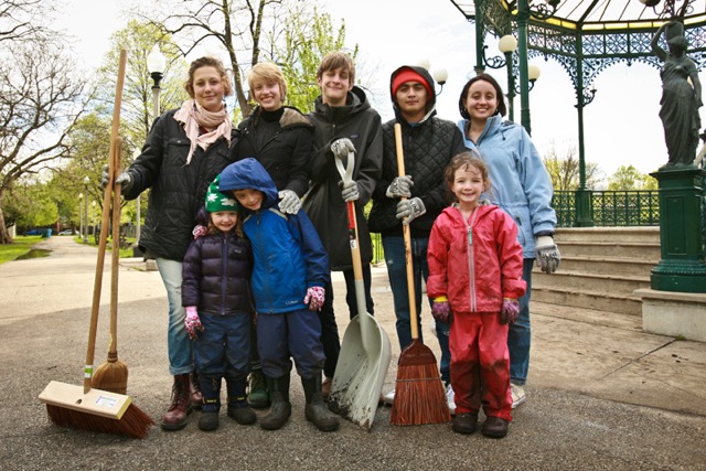 A group of people, both children and adults post for a photo outside in front of a gazebo with brooms and shovels