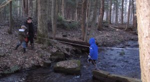 3 children crossing river in forest, wearing black and blue jackets