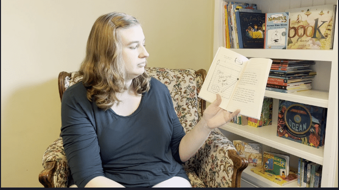 Annette Sexton reads from Timmy's monster diary in a floral chair with book case behind. Annette is a woman with blond hair, pale skin, grey shirt